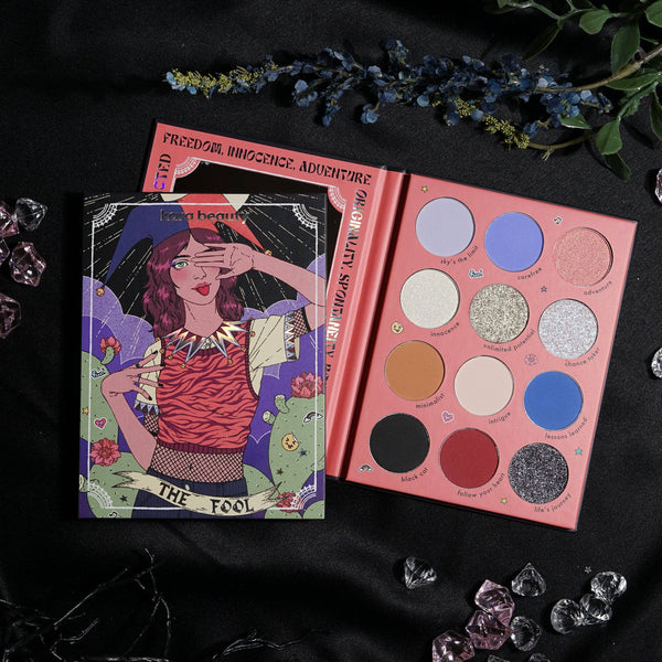 An eye palette with 12 shades including matte, shimmer, and glitter finishes, with a graphic of a woman with a tarot card in the background.