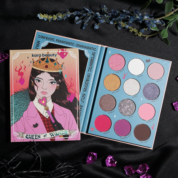 A colorful eye palette named 'QUEEN OF WANDS' with various shades of matte, shimmer, and glitter eyeshadows. The palette is open next to its packaging which features an illustration of a woman wearing a crown.